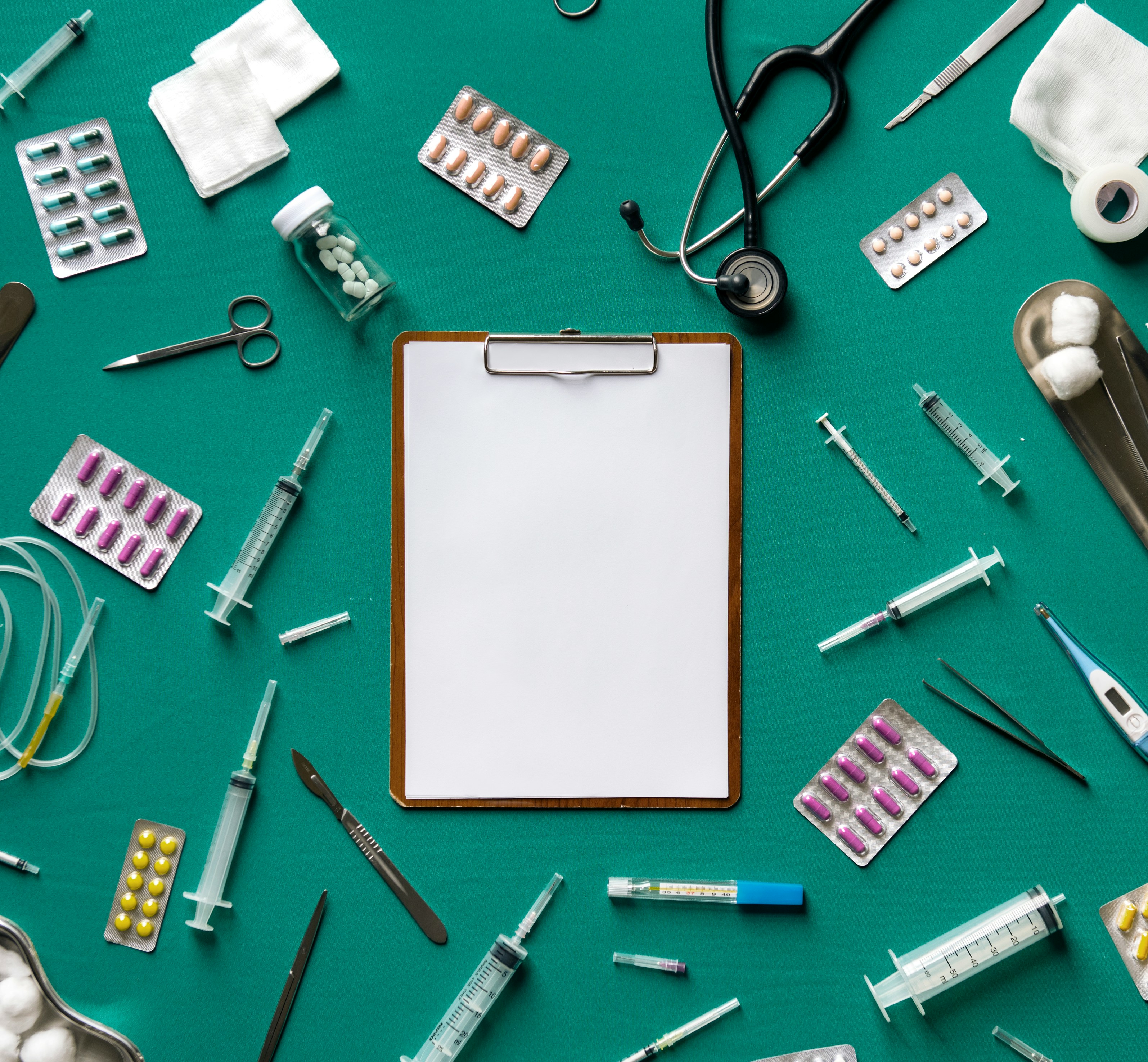 Flat-lay of clinical instruments — stethoscope, scalpel, syringes, blister packs of medication, gauze, and a blank clipboard — arranged on a teal surface